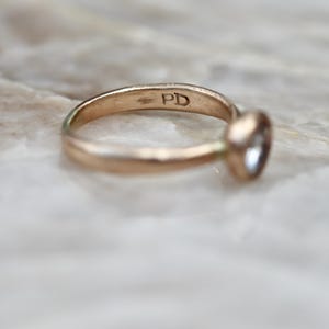 May include: A gold-toned metal ring featuring a single round bezel-set stone. The interior of the band is clearly stamped with the letters PD. The ring has a smooth, polished finish and a simple, circular band design.