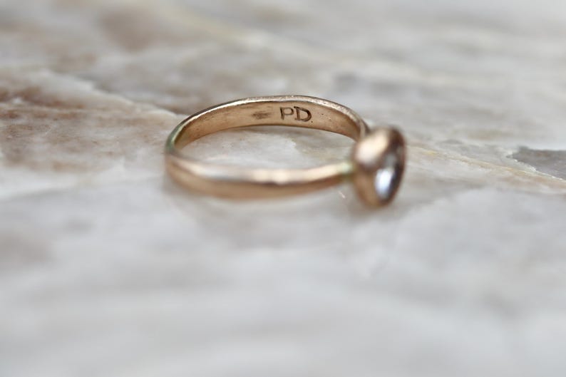 May include: A gold-toned metal ring featuring a single round bezel-set stone. The interior of the band is clearly stamped with the letters PD. The ring has a smooth, polished finish and a simple, circular band design.