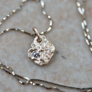 Könnte beinhalten: Eine goldfarbene Halskette mit einem Anhänger. Der Anhänger hat eine unregelmäßige, strukturierte Form und einen kleinen, hellblauen Edelstein. Die zarte Kette ist ebenfalls goldfarben, was ein elegantes Schmuckstück ergibt.