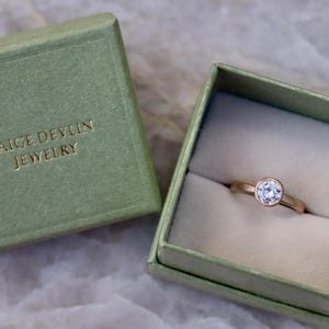 May include: A gold ring with a single round clear stone in a bezel setting, presented inside an open sage green square box. The lid of the box is placed beside it, featuring the gold embossed text PAIGE DEVLIN JEWELRY on the top surface.