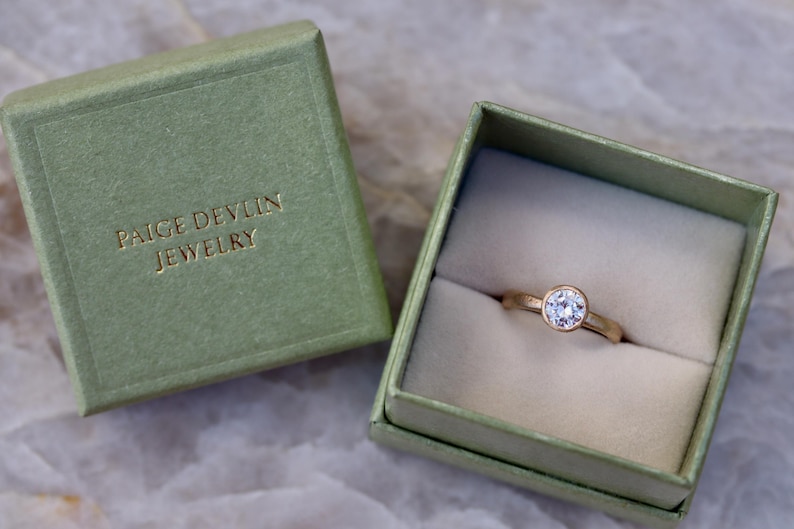 May include: A gold ring with a single round clear stone in a bezel setting, presented inside an open sage green square box. The lid of the box is placed beside it, featuring the gold embossed text PAIGE DEVLIN JEWELRY on the top surface.