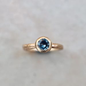 May include: A gold ring featuring a single round, faceted blue gemstone set in a bezel setting. The band of the ring has a textured, hammered finish, providing a distinct surface appearance that contrasts with the smooth, polished bezel holding the stone.