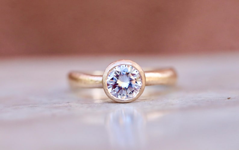 May include: A gold ring featuring a single round, clear, faceted gemstone set in a smooth bezel setting. The band has a rounded, polished finish, and the center stone displays multiple facets that reflect light with a bright, clear appearance.