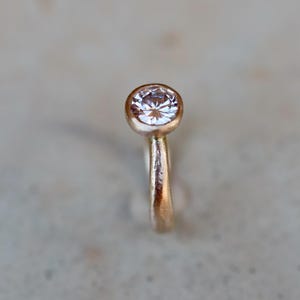 May include: A ring featuring a single round, clear faceted gemstone set in a bezel setting. The band is crafted from a textured, warm-toned metallic material with a matte finish, showcasing a simple and clean design focused on the central stone.