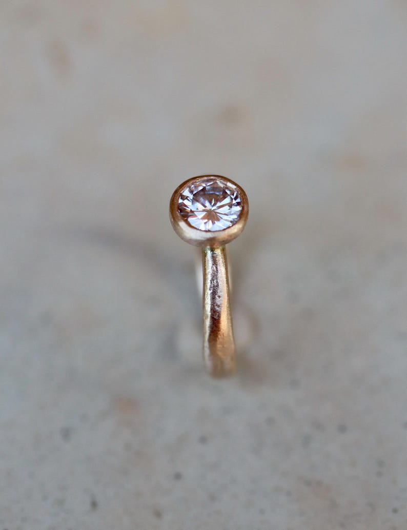 May include: A ring featuring a single round, clear faceted gemstone set in a bezel setting. The band is crafted from a textured, warm-toned metallic material with a matte finish, showcasing a simple and clean design focused on the central stone.