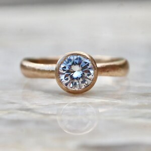 May include: A ring featuring a round, clear, faceted gemstone set in a bezel setting. The band has a matte, textured finish with a warm, metallic copper-toned appearance. The stone is positioned centrally, reflecting light with multiple facets.