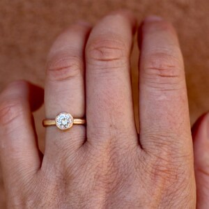 May include: A gold ring featuring a single round brilliant-cut white gemstone set in a smooth bezel setting. The band is a simple, polished gold metal. The ring is positioned on a finger, showcasing the circular stone and the gold band design.
