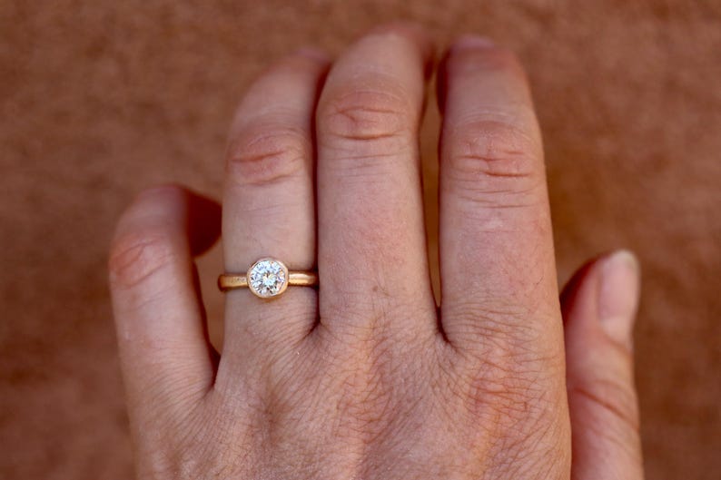 May include: A gold ring featuring a single round brilliant-cut white gemstone set in a smooth bezel setting. The band is a simple, polished gold metal. The ring is positioned on a finger, showcasing the circular stone and the gold band design.