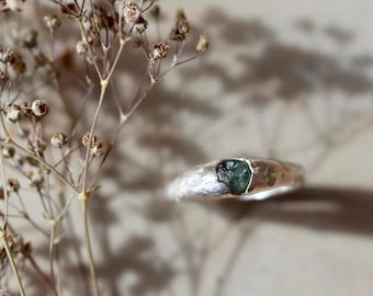 May include: A silver ring with a hammered, textured band featuring a single, small, raw, dark green gemstone set securely in the center. The metal surface has an uneven, organic finish that contrasts with the deep color of the stone.