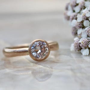 May include: A rose gold ring featuring a single round, brilliant-cut clear gemstone set in a smooth bezel setting. The band has a matte, textured finish, and the ring is positioned next to a cluster of small, white yarrow flowers.
