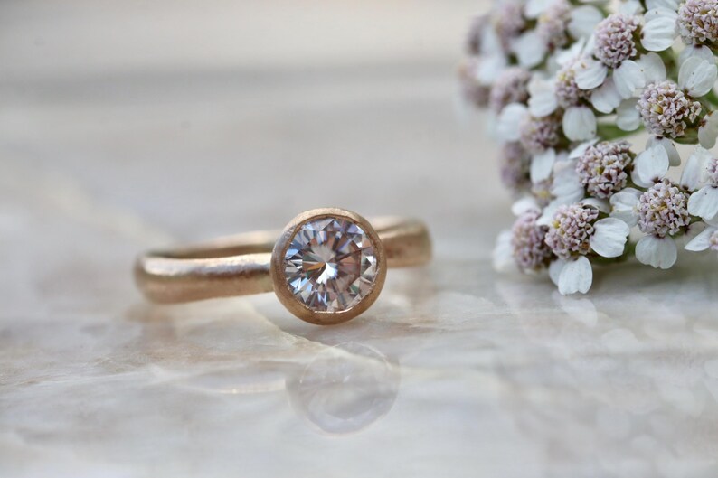 May include: A rose gold ring featuring a single round, brilliant-cut clear gemstone set in a smooth bezel setting. The band has a matte, textured finish, and the ring is positioned next to a cluster of small, white yarrow flowers.