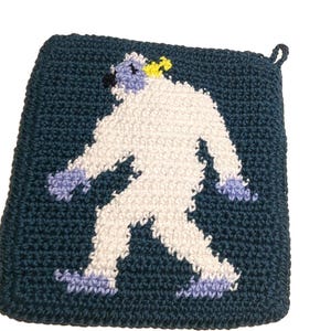 May include: A square, dark blue crocheted potholder featuring a centered, white, furry-textured creature with light purple hands and feet, a small black nose, and a tiny yellow detail on its head, complete with a small loop for hanging.
