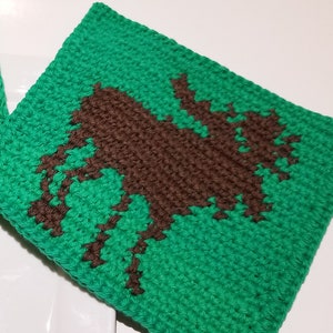 May include: Two square, green crocheted potholders with a brown moose silhouette. The potholders are placed on a white rectangular plate. The moose design is centered on each potholder.