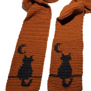 May include: A burnt orange crochet scarf featuring a repeating pattern of a black cat sitting on a horizontal line beneath a crescent moon. The scarf is constructed with a dense, textured stitch pattern and displays the cat motif on both ends.