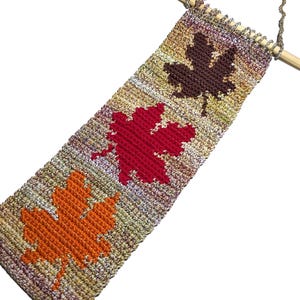 May include: A rectangular crochet wall hanging featuring three distinct maple leaf motifs in dark brown, bright red, and vibrant orange. The background is a variegated yarn in earth tones, suspended from a light wooden dowel with a metal chain.