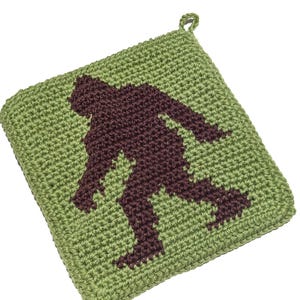 May include: A square, green crocheted potholder featuring a dark brown silhouette of Bigfoot. The potholder has a loop for hanging and is set against a white background. The Bigfoot design is centered on the potholder.