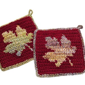 May include: Two square crochet potholders in deep burgundy yarn, each featuring a central maple leaf motif in variegated gold, yellow, and orange tones. The edges are finished with a metallic gold border and a small loop for hanging.