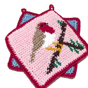 Puede incluir: Dos agarraderas de ganchillo. La agarradera superior es rosa con un borde rojo y un diseño de pájaro. El pájaro tiene la cabeza roja, el cuerpo blanco y las alas marrones. El pájaro está posado en una rama marrón con hojas verdes.