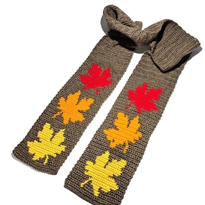 May include: A brown crocheted scarf featuring a repeating pattern of maple leaf motifs in vibrant shades of red, orange, and yellow. The scarf is constructed with a dense, textured stitch pattern and displays two distinct ends with leaf designs.