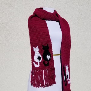 May include: A burgundy crocheted scarf with a black and white cat design. The scarf has fringe on the ends.