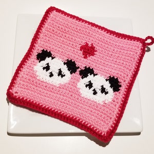 May include: A handmade, square, pink crochet potholder with a red border. The potholder features two panda faces with black and white details and a small red heart. A loop is attached for hanging.