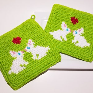 Może przedstawiać: Dwa zielone, szydełkowe uchwyty na garnki z białymi królikami i czerwonymi sercami. Króliki się całują, a serca są nad nimi.