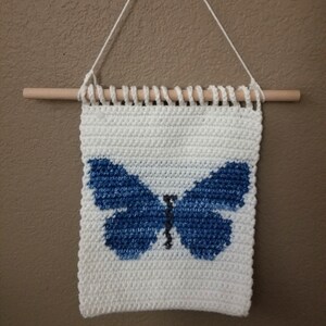 May include: A white crocheted wall hanging with a blue butterfly design. The hanging is attached to a wooden dowel rod with a white string.