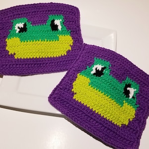 May include: Two purple crocheted squares with a green and yellow frog face design. The frog has black eyes with white pupils.