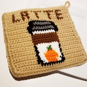 May include: A square, tan crochet potholder featuring a dark brown coffee cup graphic with a white lid and a small orange pumpkin icon. The word LATTE is stitched in dark brown letters at the top. It includes a small loop for hanging.