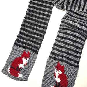 May include: A handmade crochet scarf featuring a repeating pattern of black and gray stripes. The ends of the scarf have a gray background with a red and white fox design. The scarf is made of soft yarn.