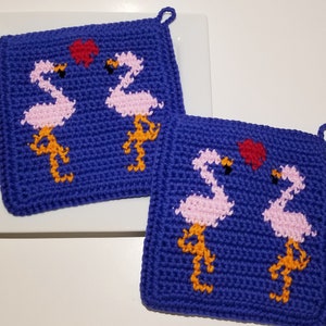 Könnte beinhalten: Zwei gehäkelte Topflappen mit blauem Hintergrund und rosa Flamingos mit orangenen Beinen. Ein rotes Herz befindet sich zwischen den Flamingos auf jedem Topflappen.