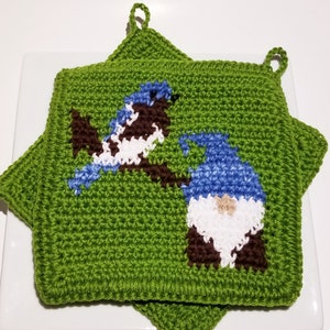Puede incluir: Dos agarraderas verdes de ganchillo con un diseño de pájaro y gnomo. El pájaro es azul, blanco y marrón, posado en una rama marrón. El gnomo tiene un gorro azul, barba blanca y cuerpo marrón. Cada agarradera tiene un lazo para colgar.