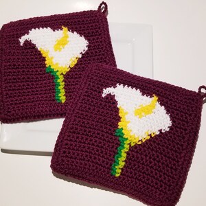 May include: Two crocheted pot holders with a white flower design. The pot holders are burgundy with green and yellow accents.