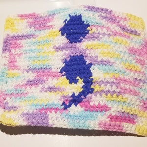 Può includere: Un quadrato all'uncinetto nei colori pastello rosa, viola, giallo e blu, con un grande punto e virgola blu al centro. L'articolo fatto a mano è probabilmente un pezzo decorativo o una piccola coperta.