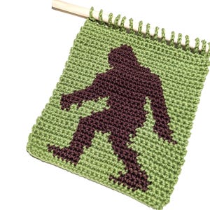 Op de afbeelding: Een groen en bruin gehaakte wandhanger met een silhouet van Bigfoot. De Bigfoot-figuur is donkerbruin tegen een groene achtergrond. De wandhanger is bevestigd aan een houten deuvel.