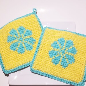 May include: Two square crochet potholders. Each potholder features a yellow background with a turquoise flower design in the center. The edges are trimmed with turquoise yarn, and a small loop is attached for hanging.
