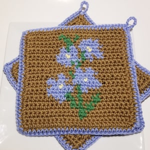 Puede incluir: Dos agarraderas de olla de color marrón hechas a ganchillo con un borde azul y un diseño floral en el centro. Las flores son azules con centros amarillos y hojas verdes.