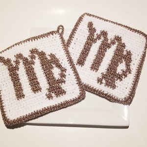 May include: Two square crochet potholders featuring a white background with a brown Virgo zodiac symbol stitched in the centre. Each potholder has a brown crocheted border and a small loop at the corner for hanging, showcasing a textured yarn finish.