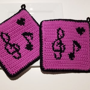 May include: Two pink crocheted pot holders with black trim and musical notes. The pot holders have a loop for hanging.
