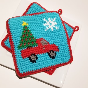 May include: Two crocheted pot holders with a Christmas theme. One pot holder features a red truck carrying a green Christmas tree with a yellow star on top. The other pot holder has a white snowflake design. Both pot holders have a blue background and a red border.