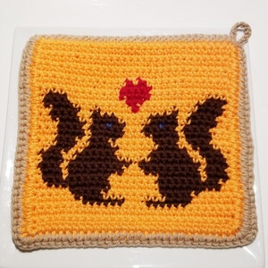 Könnte beinhalten: Ein gehäkelter Topflappen mit gelbem Hintergrund und zwei braunen Eichhörnchen, die ein rotes Herz halten. Der Topflappen hat einen braunen Rand.