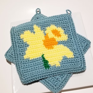May include: Two crocheted pot holders with a star shape. The top pot holder features a yellow daffodil with an orange center and green leaves on a blue background.
