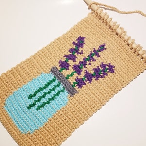 May include: Crocheted wall hanging featuring a blue mason jar with green stems and purple flowers. The hanging is beige and has a wooden dowel at the top.