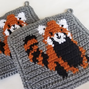 Puede incluir: Dos agarraderas de olla de crochet gris con diseños de panda rojo. Los pandas rojos están hechos a crochet en hilo naranja, negro y blanco.