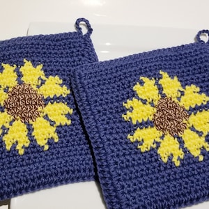 Può includere: Due presine da forno in crochet blu con motivi di girasole gialli e marroni.
