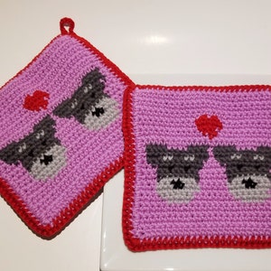May include: Two pink crocheted pot holders with red borders and a heart design. Each pot holder features two grey crocheted dogs with black eyes.