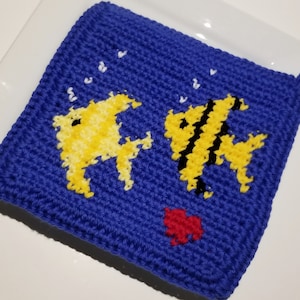 May include: A square, blue crocheted potholder featuring two yellow fish with black stripes and accents. The fish are swimming near white bubbles and a small red heart. A loop is attached for hanging.