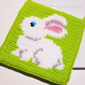 Può includere: Un sottopentola all'uncinetto verde con un design di coniglio all'uncinetto bianco. Il coniglio ha un orecchio rosa e grigio e occhi blu.