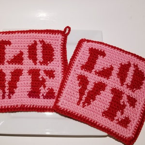 Può includere: Due presine da forno in crochet rosa e rosso con la parola "LOVE" scritta in rosso.