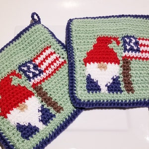 Op de afbeelding: Twee groene gehaakte ovenwanten met een blauwe rand. Elke ovenwant heeft een gnome met een rode muts en een witte baard die een Amerikaanse vlag vasthoudt.